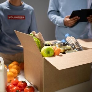 People packing a cardboard box with essentials like fruits, vegetables, and bottled water for charity.